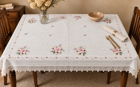 A square white tablecloth with a delicate lace border and embroidered pink rose motifs, styled on a wooden dining table with a vase of dried flowers, a wooden bowl, and rolled napkins.
