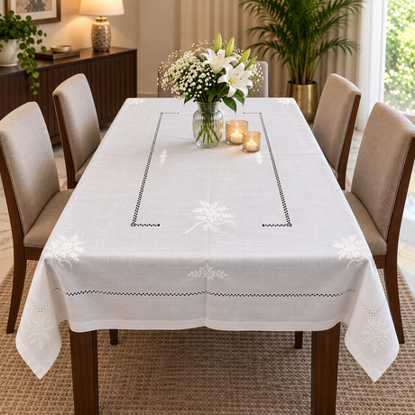 A white embroidered tablecloth with delicate floral and geometric patterns draped over a wooden dining table, surrounded by six upholstered chairs in a bright, modern dining room.