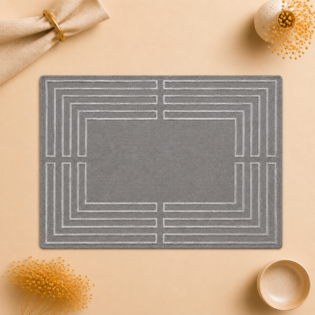 A rectangular gray placemat with a white geometric border pattern, styled in a flat-lay on a beige background with decorative dried flowers and a napkin.