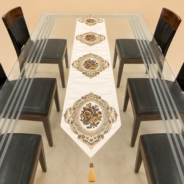 An ornate white table runner with four matching oval medallions and a tassel, displayed on a glass-top dining table surrounded by six dark chairs.