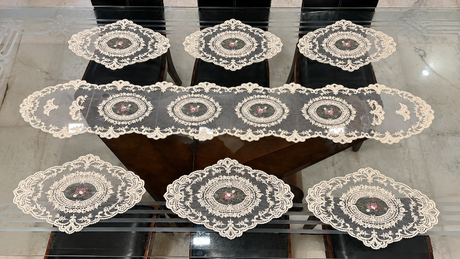 A set of seven white lace table linens, including a long runner and six matching placemats, arranged on a glass-topped table over dark chairs.