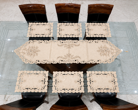 A dining table set with a long cream lace table runner and six matching square placemats, surrounded by dark wooden chairs on a light marble-patterned floor.