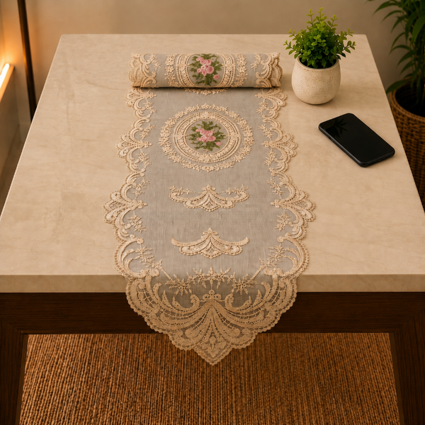 An ornate beige lace table runner with a floral embroidered medallion is draped over a light beige stone table, with a matching runner rolled up at the head. A small potted plant and a smartphone sit on the table.