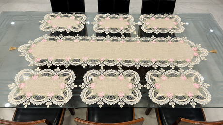 A set of beige table linens including a long runner and six matching placemats with white floral lace trim and pink rose accents, arranged on a glass-top dining table with chairs visible.