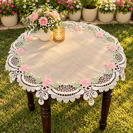A round lace tablecloth with pink floral embroidery and green leaf accents, draped over a dark wooden table in a sunny garden setting.