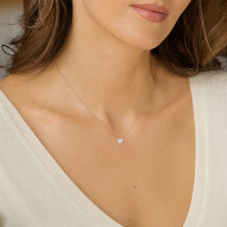 A close-up of a woman wearing a delicate gold chain necklace with a small heart-shaped clear gemstone pendant, styled with a cream-colored V-neck top.