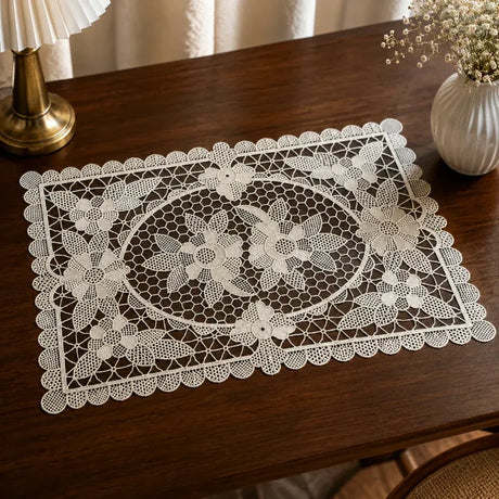 A white lace placemat with an intricate floral pattern and scalloped border, displayed on a dark wooden table next to a lamp and a vase of flowers.
