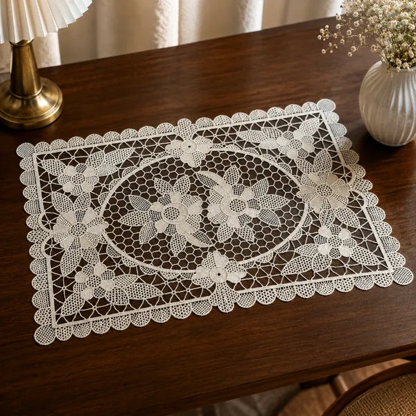 A white lace placemat with an intricate floral pattern and scalloped border, displayed on a dark wooden table next to a lamp and a vase of flowers.