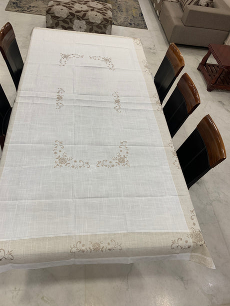 A white tablecloth with beige embroidered floral patterns and a matching beige border, draped over a dining table surrounded by dark wooden chairs in a home setting.