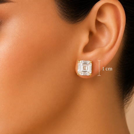 Close-up of a model's ear wearing a square clear gemstone stud earring with a gold setting, with a '1 cm' size marker shown next to it.
