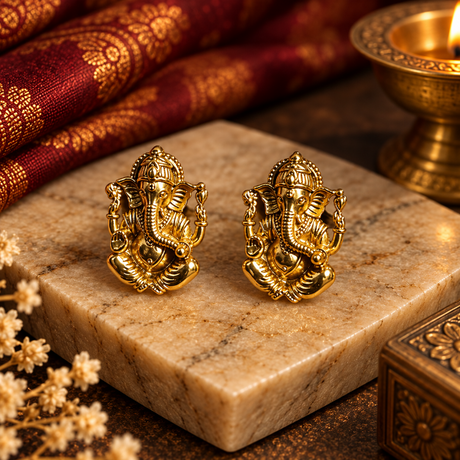 A pair of gold-toned Ganesha stud earrings displayed on a rectangular marble slab, with a lit brass oil lamp and ornate red fabric in the background.