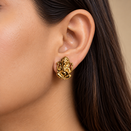 A close-up of a woman's ear wearing a small gold Ganesha stud earring against a soft beige background.