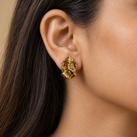 A close-up of a woman's ear wearing a textured gold Ganesha-shaped stud earring, set against a soft beige background.