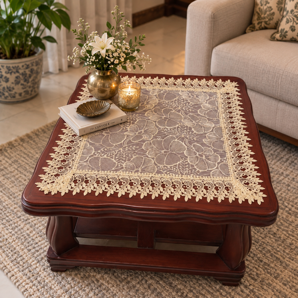 A square dark wood coffee table in a living room setting, covered with a beige floral lace tablecloth. Decorative items on top include a gold vase with white flowers, a candle, a small bowl, and a book.
