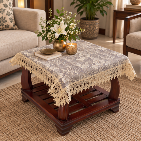 A square lace tablecloth with a floral pattern and cream crochet trim, draped over a dark wood coffee table in a cozy living room setting.