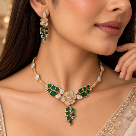 A close-up lifestyle shot of a model wearing a gold and green leafy floral necklace and matching drop earrings. The jewelry features emerald-cut green stones and brilliant-cut clear stones set in gold, shown on a model in a beige embroidered garment.