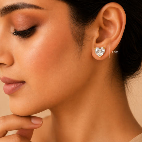 A close-up of a woman's ear wearing a small heart-shaped stud earring. The earring appears silver-toned with a clear gemstone, and a small '1 cm' size marker is shown beside it.