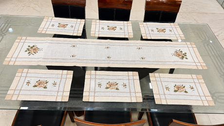 A set of six table linens including one long runner and five matching placemats, laid out on a glass-top dining table with dark wooden chairs. The linens are cream-colored with beige stripes and embroidered floral motifs.