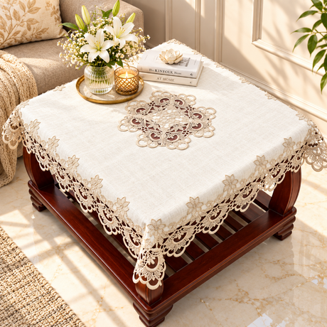 An off-white lace tablecloth with floral embroidery and a central medallion design, draped over a dark wood coffee table in a bright, modern living room setting.