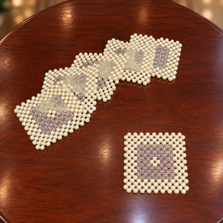 A set of square beaded coasters made of cream-colored beads with gray and white geometric patterns, displayed on a dark brown wooden table.