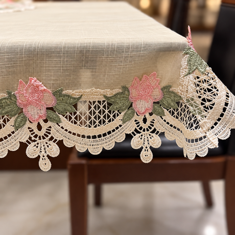 A beige tablecloth with a decorative scalloped lace border and embroidered pink flowers with green leaves, draped over a wooden chair in an indoor setting.