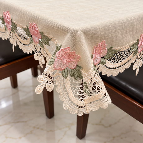 Beige tablecloth with pink floral embroidery and lace trim draped over a dining table, shown in a home setting with dark wooden chairs and a tiled floor.