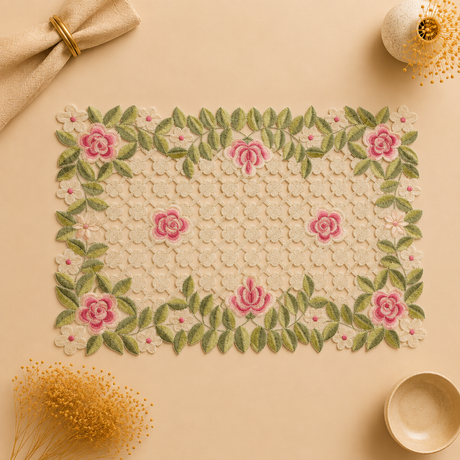 An embroidered rectangular placemat with a floral border and central pattern, styled on a beige surface with dried flowers, a napkin, and a small bowl.