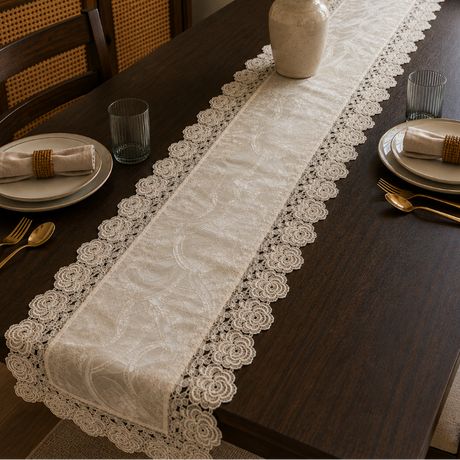 A cream lace table runner with floral embroidery and scalloped edges laid down the center of a dark wood dining table. The table is styled with place settings, gold cutlery, a vase of white flowers, and chairs in a warm, well-lit dining room.