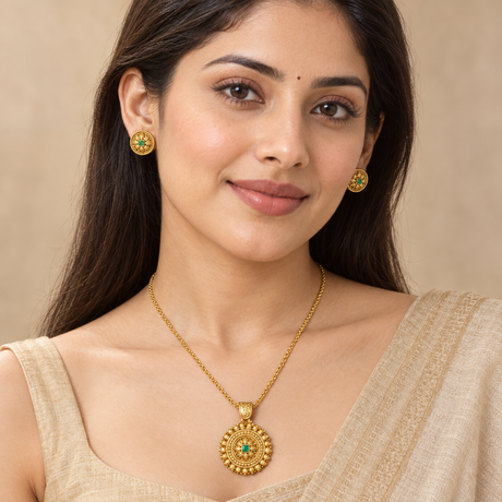 A woman wearing a gold necklace with a round pendant and matching gold stud earrings, each featuring a green stone accent, against a soft beige background.