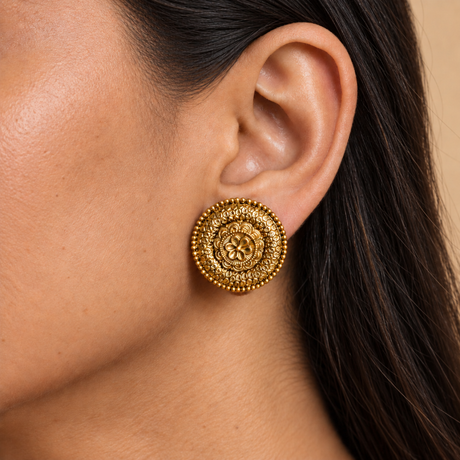 A close-up of a woman's ear wearing a round, ornate gold earring with intricate filigree detailing and a beaded border.