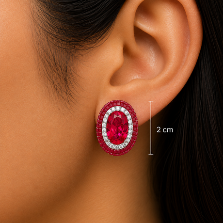 Close-up of a model's ear wearing an oval stud earring with a large red center stone and surrounding clear stones, with a vertical measurement marker indicating 2 cm width.