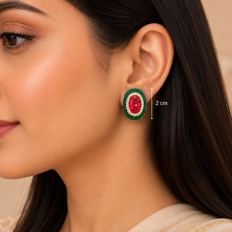 A close-up of a woman's ear wearing an oval stud earring with a red center, green outer ring, and gold-toned detailing. A vertical measurement marker indicates the earring is approximately 2 cm wide.
