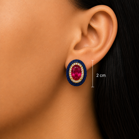 Close-up of a model's ear wearing an oval stud earring with a deep blue beaded border, a gold-toned inner ring, and a central oval pink gemstone. A vertical measurement marker reads 2 cm.