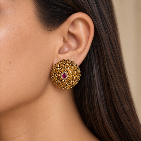 A close-up of a woman's ear wearing a large, ornate gold-toned Indian-style earring with a central pink gemstone.