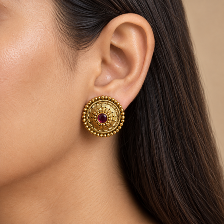 A close-up of a model's ear wearing a round, ornate gold-toned Indian-style stud earring with a central red stone.