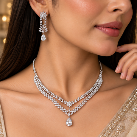 A close-up lifestyle shot of a woman wearing a sparkling silver-tone double-row necklace with a teardrop pendant and a matching drop earring. The jewelry is set with clear gemstones, shown on a model in a beige embroidered garment.