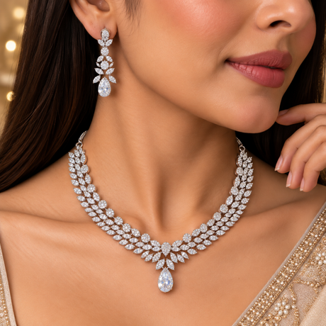 A close-up lifestyle shot of a woman wearing a sparkling diamond necklace and matching drop earrings. The jewelry features intricate silver-toned metal settings with numerous clear gemstones, including a teardrop-shaped center stone on the necklace.
