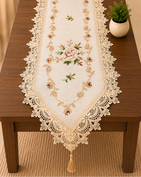 An embroidered table runner with a delicate lace border and tassel, displayed on a dark wooden dining table next to a small potted green plant.