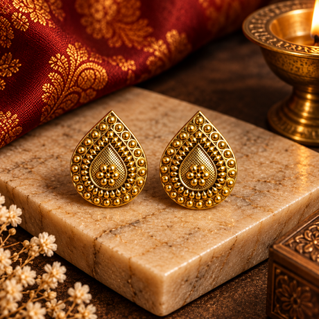 A pair of ornate gold teardrop-shaped earrings with intricate beadwork and floral motifs, displayed on a beige marble block with a red patterned fabric and a brass oil lamp in the background.