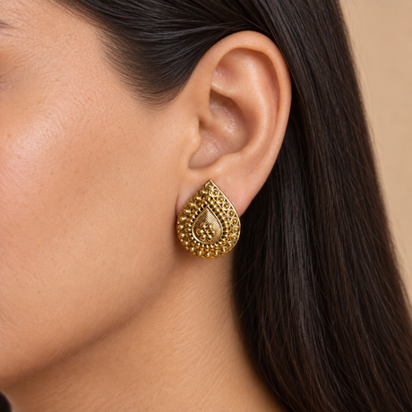 A close-up of a model's ear wearing a teardrop-shaped gold earring with intricate textured detailing.