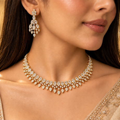 A close-up of a woman wearing a gold-toned choker necklace and matching drop earring, both heavily set with clear stones. The jewelry is shown on a model wearing a beige embellished top against a warm, softly lit background.