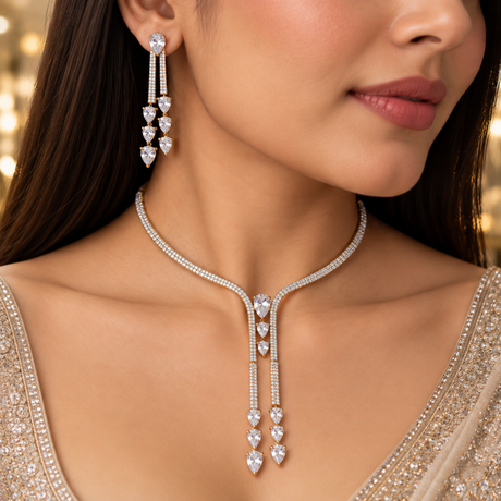 A close-up lifestyle shot of a woman wearing a sparkling silver-toned Y-shaped necklace and matching drop earrings adorned with clear heart-shaped gemstones, set against a softly blurred warm background.