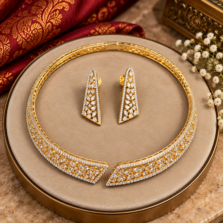 Gold-toned jewelry set with a wide collar-style necklace and matching earrings, both heavily embellished with clear stones, displayed on a round beige jewelry box. Red patterned fabric and small white flowers decorate the scene.