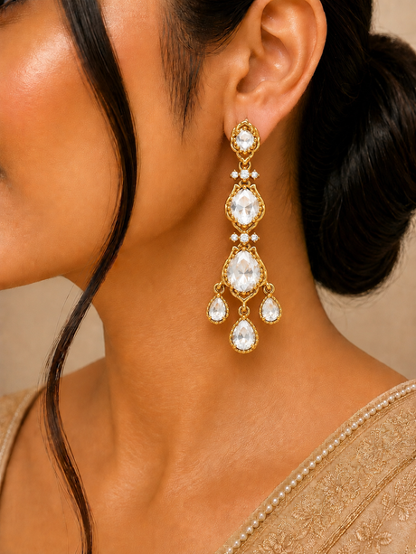 Close-up of a woman wearing gold earrings with clear stones, against a neutral background.