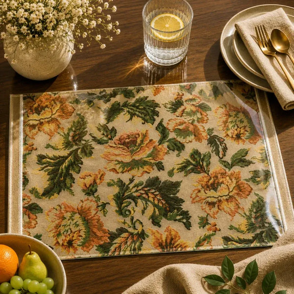 A rectangular woven placemat with a vintage floral pattern in green, orange, and beige, styled on a dark wooden dining table with a glass of lemon water, plates, cutlery, and a vase of baby's breath.