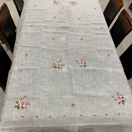 A white woven tablecloth with embroidered pink roses and green leaves, draped over a rectangular dining table surrounded by dark wooden chairs.