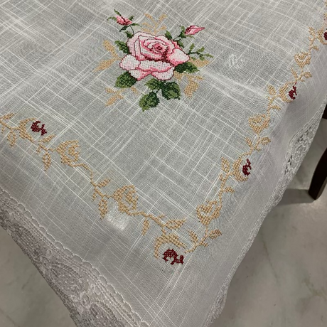 A white woven tablecloth with a delicate pink rose and green leaves embroidered in the center, finished with a gold floral border and lace trim along the edges.