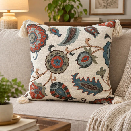 A square decorative throw pillow with a cream background and a colorful floral and paisley pattern in muted blue, rust red, and taupe, styled on a beige sofa in a cozy living room setting.