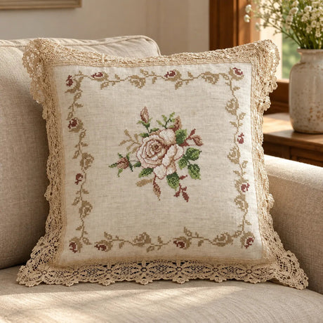 A square decorative throw pillow with a floral cross-stitch design and lace trim, resting on a beige sofa in a warm, sunlit room.
