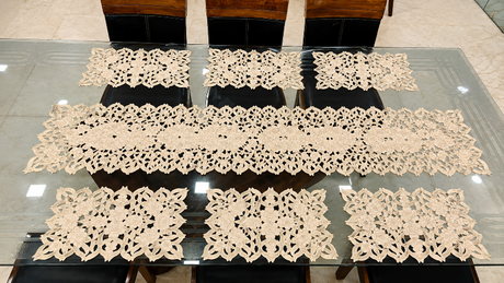 A set of cream-colored crocheted table runners and matching placemats arranged on a glass-top dining table with dark chairs.